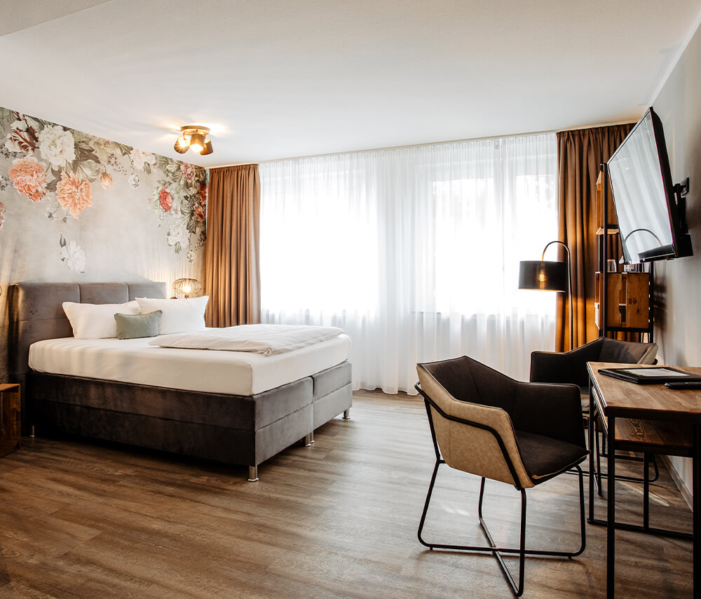 Elegantes Zweibettzimmer im Hotel Westerkamp in Osnabrück, ausgestattet mit einem großen Doppelbett mit grauem Kopfteil und weißer Bettwäsche. Die Wand hinter dem Bett ziert eine große, blumige Tapete mit zarten Pastelltönen. Natürliches Licht flutet durch die bodentiefen Fenster, die mit weißen, transparenten Vorhängen und braunen Schals ausgestattet sind. Das Zimmer bietet eine gemütliche Sitzecke mit zwei modernen, braunen Stühlen und einem kleinen Tisch sowie einen an der Wand montierten Flachbildfernseher. Der Holzboden verleiht dem Raum eine warme Atmosphäre.