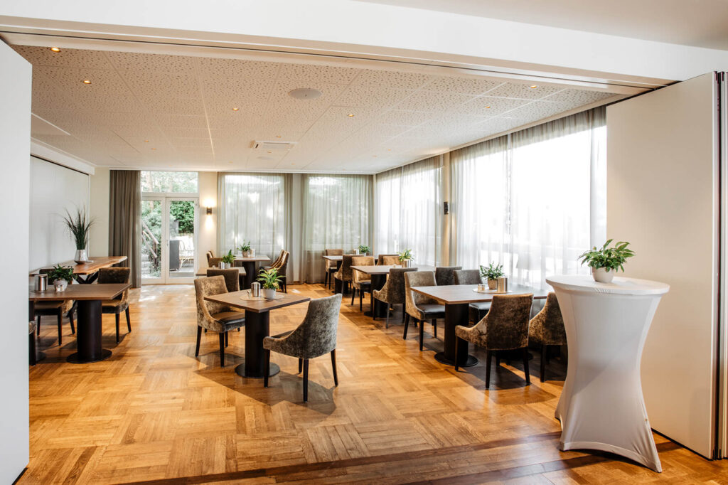 Der elegante und helle Speiseraum, der auch für Feiern benutzt werden kann, des Hotel Westerkamp in Osnabrück. Der Raum ist mit einem warmen Parkettboden ausgelegt und durch große Fenster, die reichlich Tageslicht hereinlassen, beleuchtet. Mehrere Tische mit stilvollen, dunklen Holzplatten und bequemen, samtbezogenen Stühlen sind im Raum verteilt. An den Tischen stehen kleine, gepflegte Grünpflanzen, die für eine frische und natürliche Atmosphäre sorgen. Im Hintergrund öffnet sich eine Tür zu einem angrenzenden Raum oder Garten, der den Gästen eine ansprechende Aussicht bietet.