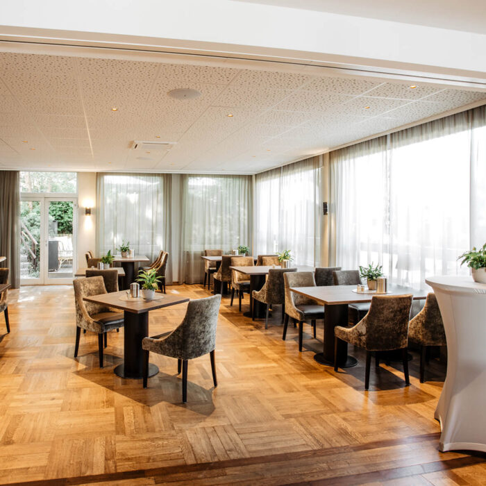 Der elegante und helle Speiseraum, der auch für Feiern benutzt werden kann, des Hotel Westerkamp in Osnabrück. Der Raum ist mit einem warmen Parkettboden ausgelegt und durch große Fenster, die reichlich Tageslicht hereinlassen, beleuchtet. Mehrere Tische mit stilvollen, dunklen Holzplatten und bequemen, samtbezogenen Stühlen sind im Raum verteilt. An den Tischen stehen kleine, gepflegte Grünpflanzen, die für eine frische und natürliche Atmosphäre sorgen. Im Hintergrund öffnet sich eine Tür zu einem angrenzenden Raum oder Garten, der den Gästen eine ansprechende Aussicht bietet.