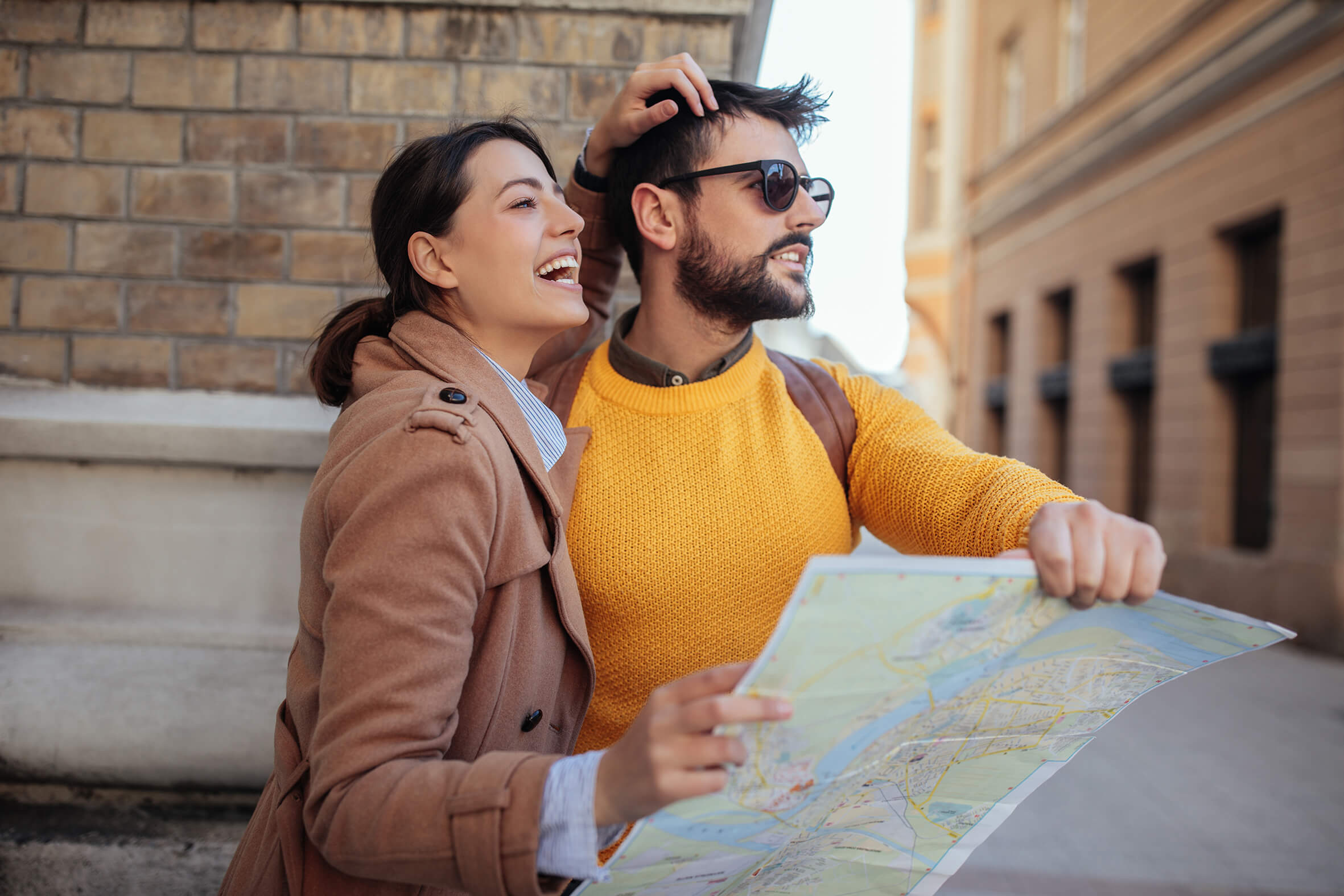 Fröhliches Paar auf Stadterkundung. Eine Frau im braunen Mantel lacht und lehnt sich an einen Mann mit einem sonnengelben Pullover und Sonnenbrille, der eine Karte hält.