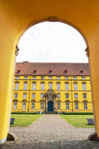 Blick auf das Schloss Osnabrück durch einen großen gelben Torbogen, der zu einem gepflasterten Innenhof führt. Das imposante Schlossgebäude besitzt drei Stockwerke und ist in einem leuchtenden Gelb gestrichen, mit zahlreichen Fenstern, die symmetrisch angeordnet sind. Über dem zentralen Eingang befindet sich ein aufwendiges Wappen, das von einem barocken Steinrahmen umgeben ist.