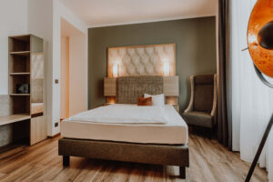 Modern eingerichtetes Zimmer im Hotel Westerkamp in Osnabrück mit einem komfortablen Bett, dessen Kopfteil mit einem stilvollen geometrischen Muster verziert ist. Links vom Bett befindet sich ein offenes Regal, das in eine Nische integriert ist, und ein langer Schreibtisch, der sich an der Wand entlangzieht. Rechts vom Bett steht ein gemütlicher, hoher Sessel neben einem eleganten Stehleuchter mit einem großen, runden, orangefarbenen Lampenschirm. Die Wände sind in einem beruhigenden Grünton gehalten, der mit der warmen Holzoptik des Bodens und der Möbel harmoniert, während die sanfte Beleuchtung eine einladende und entspannende Atmosphäre schafft.