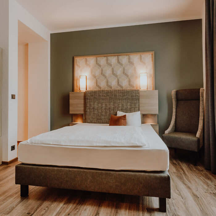Modern eingerichtetes Zimmer im Hotel Westerkamp in Osnabrück mit einem komfortablen Bett, dessen Kopfteil mit einem stilvollen geometrischen Muster verziert ist. Links vom Bett befindet sich ein offenes Regal, das in eine Nische integriert ist, und ein langer Schreibtisch, der sich an der Wand entlangzieht. Rechts vom Bett steht ein gemütlicher, hoher Sessel neben einem eleganten Stehleuchter mit einem großen, runden, orangefarbenen Lampenschirm. Die Wände sind in einem beruhigenden Grünton gehalten, der mit der warmen Holzoptik des Bodens und der Möbel harmoniert, während die sanfte Beleuchtung eine einladende und entspannende Atmosphäre schafft.