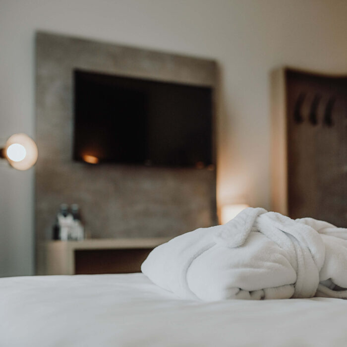 Gemütliches Zimmer im Hotel Westerkamp in Osnabrück mit einem Fokus auf ein einladendes Bett, auf dem ein weißer Bademantel gefaltet ist. Im Hintergrund ist ein moderner Flachbildfernseher an einer strukturierten Betonwand angebracht. Stilvolle Wandlampen sorgen für eine warme und einladende Atmosphäre im Zimmer.