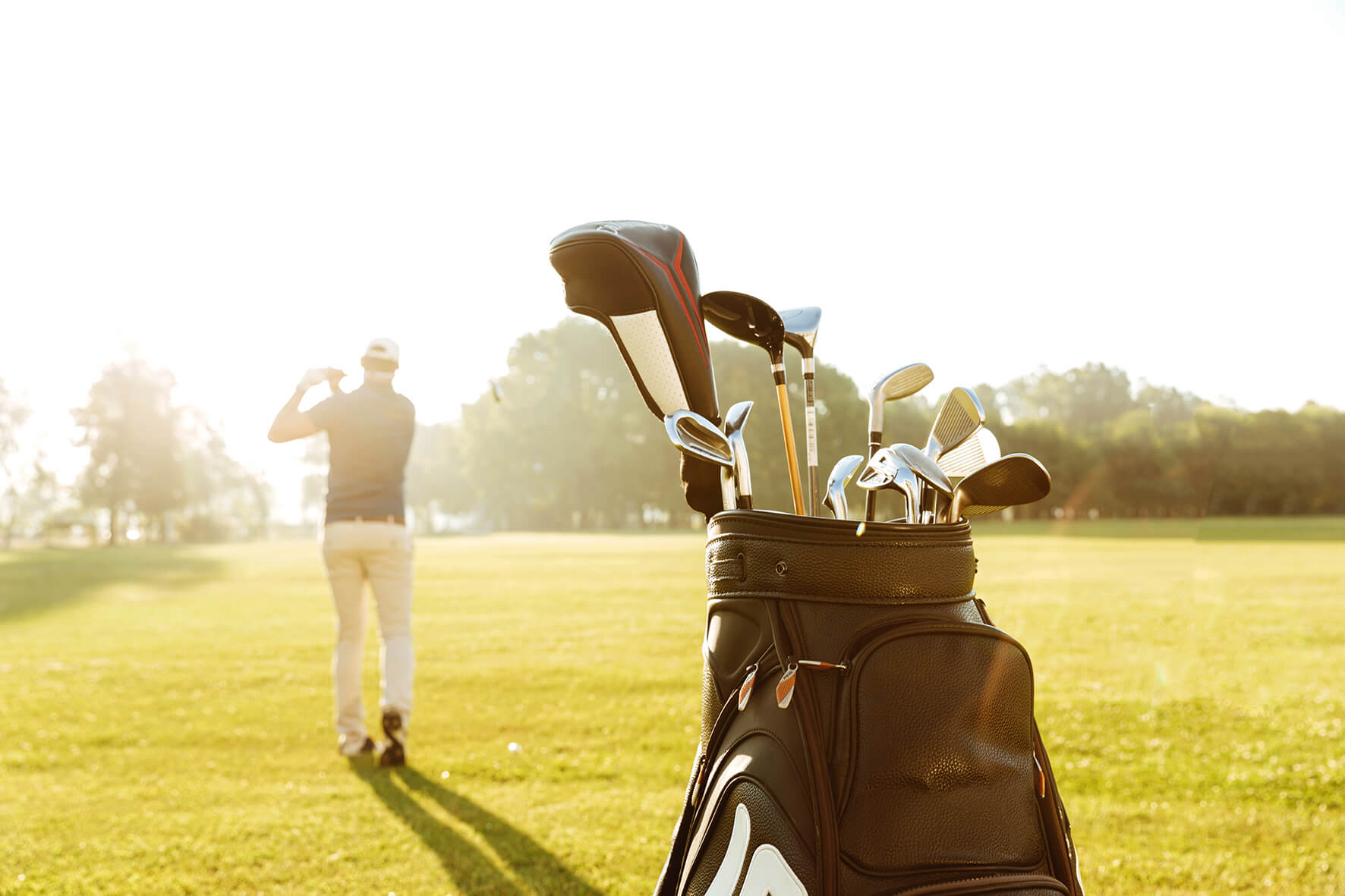 Das Bild zeigt eine Golftasche im Vordergrund, gefüllt mit einer Auswahl an Schlägern, auf einem sonnendurchfluteten Golfplatz. Im Hintergrund steht ein Golfer in einer entspannten Pose, der gerade seinen Schlag beobachtet. Er ist bekleidet mit einer Kappe, einem dunklen Poloshirt und hellen Hosen, was auf eine klassische Golfbekleidung hindeutet. Der Golfer scheint in die Ferne zu schauen, möglicherweise verfolgt er den Flug des Golfballs. Die Szene ist in warmes, goldenes Licht getaucht. Der Hintergrund ist weich fokussiert und zeigt die weite, gepflegte Rasenfläche des Golfplatzes, umgeben von einigen Bäumen am Horizont.